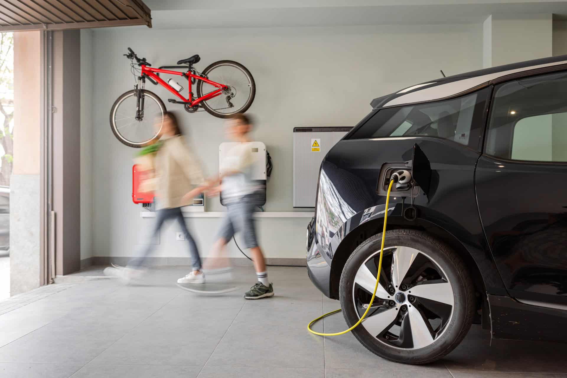 Electric vehicle charging station in a private home with an unrecognizable blurred family leaving the house, with a bicycle hanging on the wall.